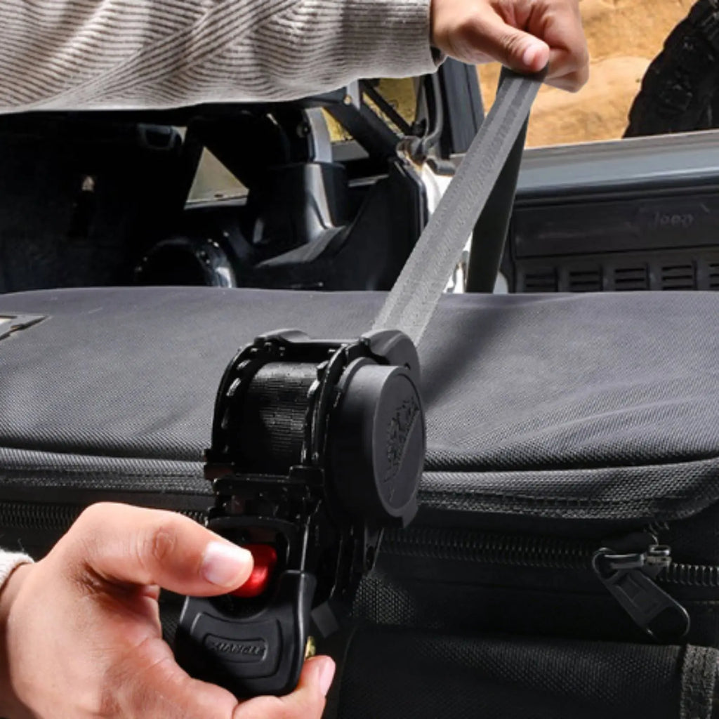 A close-up of a black car seat belt buckle attachment being used to fasten a gray seat belt to a black vehicle seat or bag, with a person's hands and arm visible in a real-world outdoor setting.