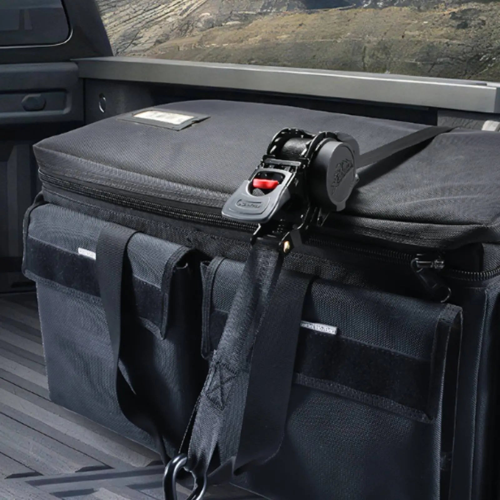 A black soft-sided utility bag or cooler with a heavy-duty strap and buckle attachment, secured inside the bed of a truck. The bag has multiple zippered compartments and side pockets, with a visible buckle strap system across the top.