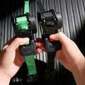 Two ratchet straps, one green and one black, held by hands against a dark background.